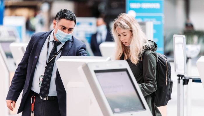 CLT Airport employee helping to service passenger on airline check-in kiosk; employee wearing face mask, passenger wearing backpack 