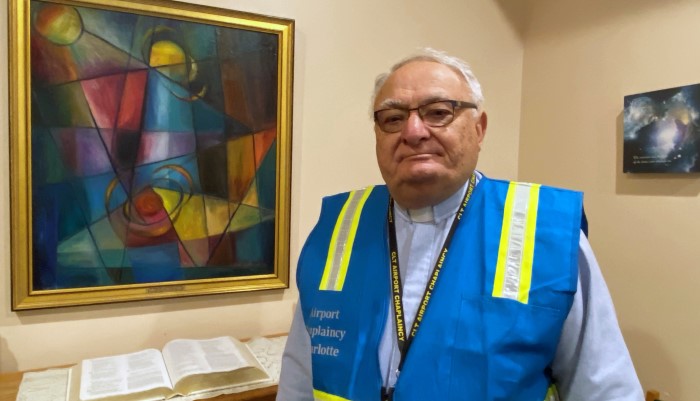 CLT Airport Chaplin featured in airport's chapel wearing safety vest and smiling, behind him is an open scripture book and colorful painting on the wall