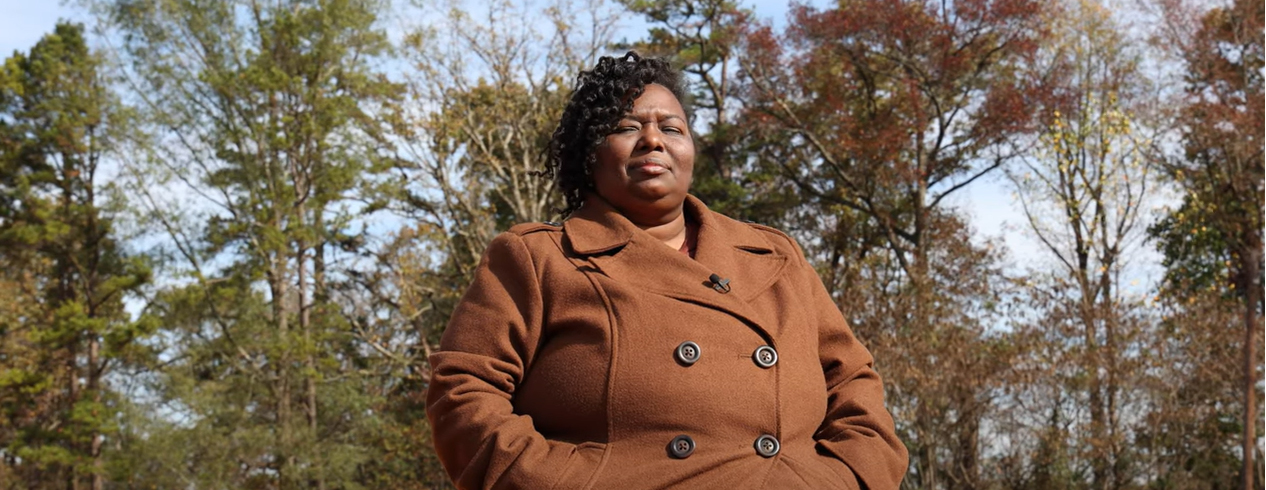 Lapri Holmes looking down towards camera wearing a brown coat with large buttons with her hands in her pockets; in the background are trees in the fall season transition