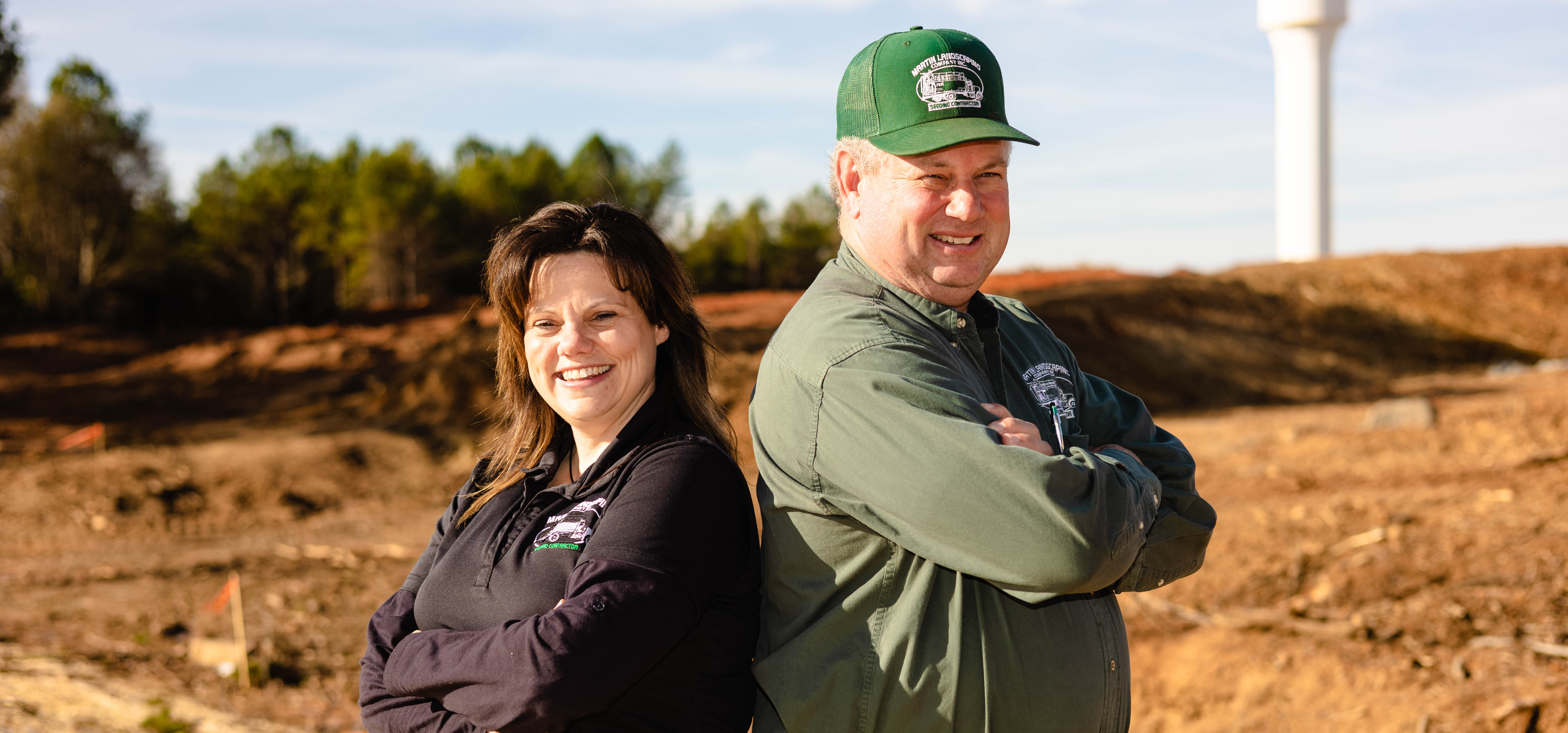 Lisa Markus (left) and David Martin (right) pose back to back with their arms crossed smiling big, behind them is a dirt field and the CLT FAA Tower miles away