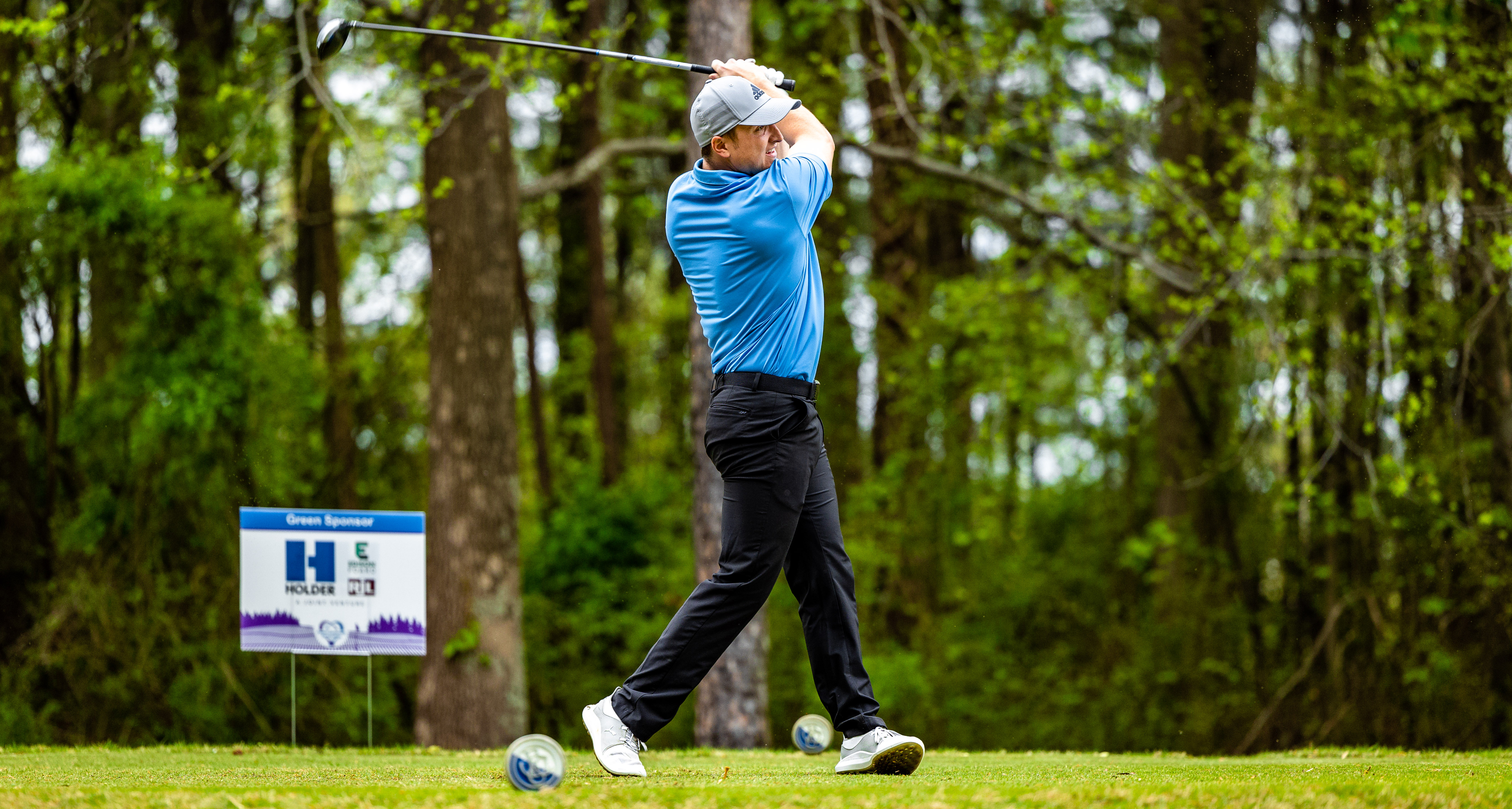 Golfer in full body swing motion, wearing blue shirt and black pants, green woods in the background