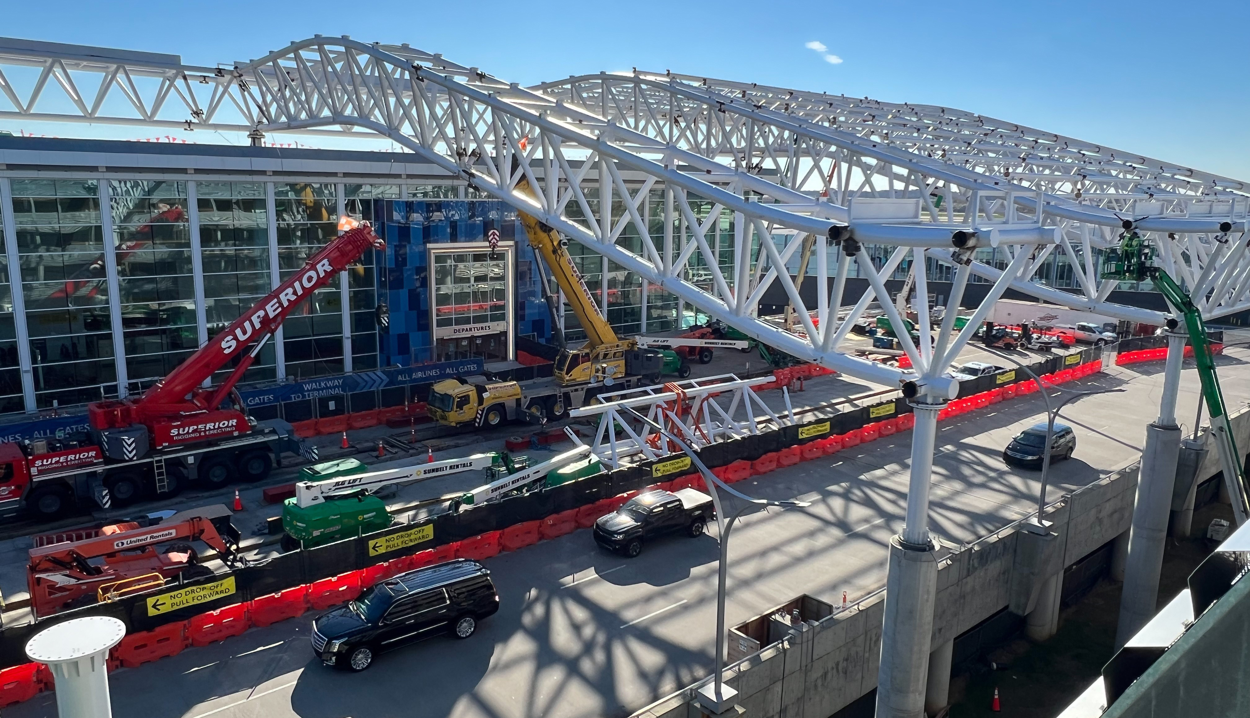 Cars make their past the front of the terminal where construction barricades and cranes sit in front of the building. In the foreground the white trusses that frame the renovation are being installed.