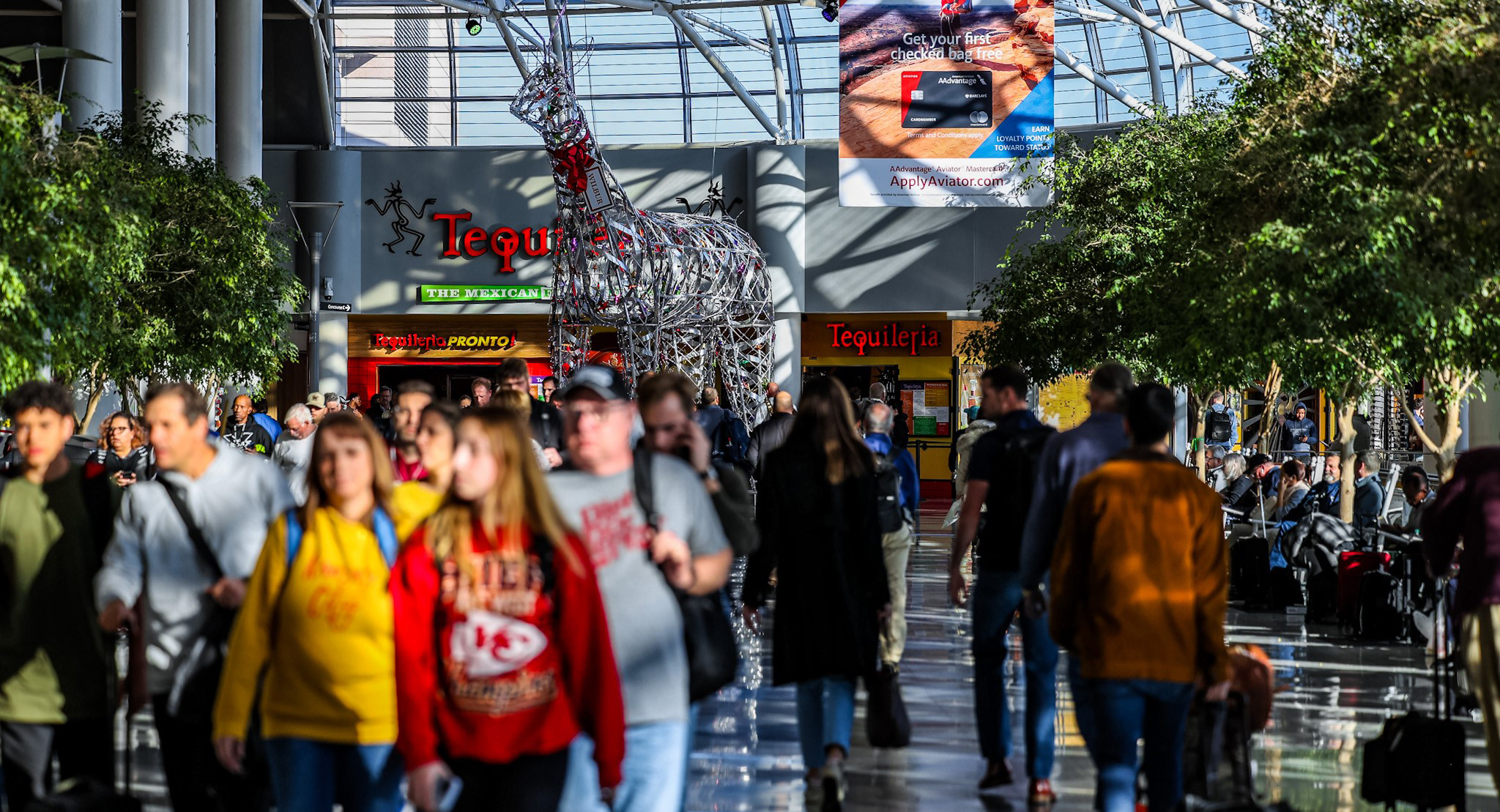 Passengers make their way through the Airport atrium where live trees line either side of the walkway. In the background is a large sculpture of a reindeer. 